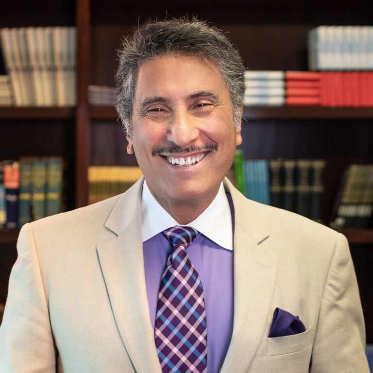 headshot of Dr. Youssef smiling at the camera in front of a bookshelf wearing a tan suit and purple shirt