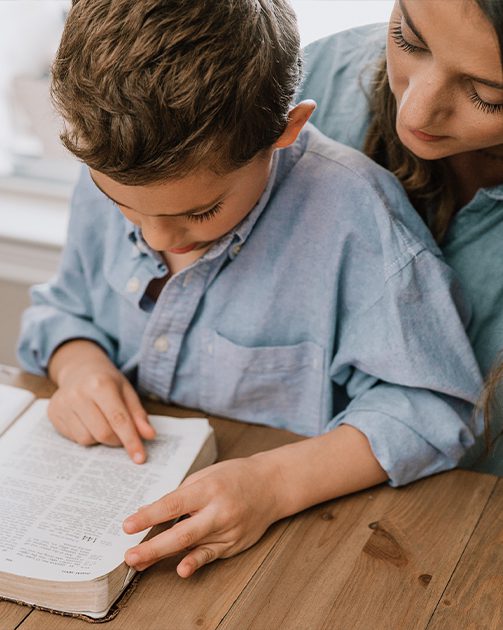 Mom smiling and helping young boy read a Bible