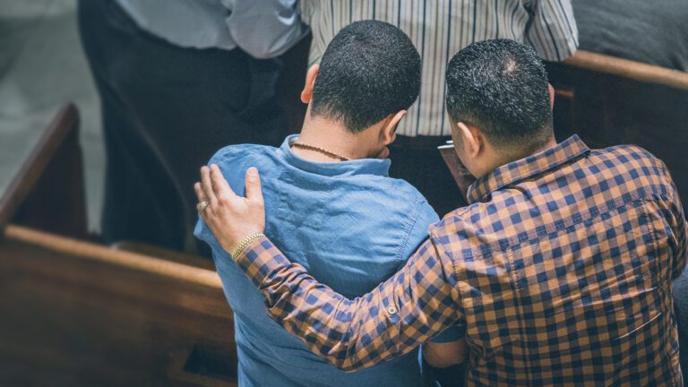 a photo of two men in a church pew, with one man's arm around the other and both praying