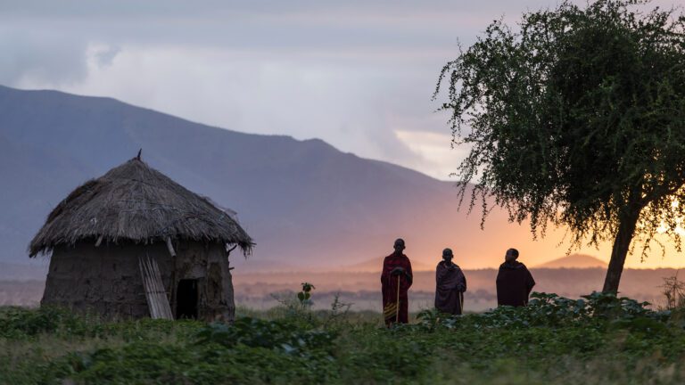 wide shot of African house, with two men and a tree nearby and mountains in the background