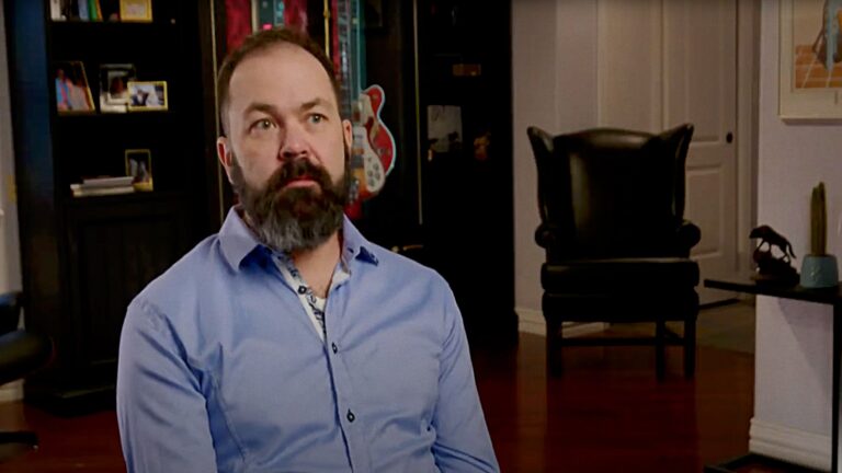 A man in a blue shirt sits in front of a bookcase